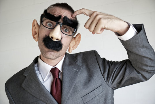 Confused businessman wearing a Groucho Marx disguise of glasses with thick eyebrows and mustache scratching his head looking at the camera