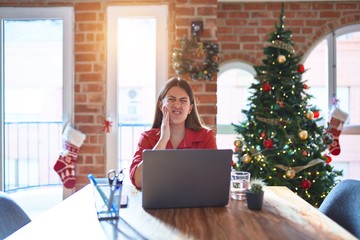 Beautiful woman sitting at the table working with laptop at home around christmas tree touching mouth with hand with painful expression because of toothache or dental illness on teeth