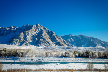 Majestic panoramic view of the valley covered with snow, turquoise river on the background of snow-capped mountains and clear blue sky. Sunny winter day in the mountains.