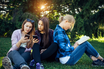 Three cute girls relax and socialize on the lawn in the summer park. Young women on the green grass among the trees, looking at smartphone, reading a book. Students in between classes.