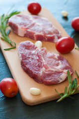Raw juicy pork steaks with red tomatoes, rosemary and garlic on a cutting board.