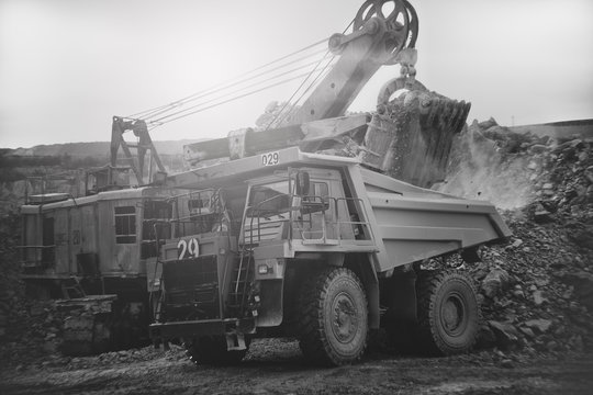 Excavator Loads Ore In The Dump-body Truck In A Quarry, Close-up. Imitation Of A Film Black And White Photography, Vignetting, Blur, Overexposed. Mining Industry. Mine And Quarry Equipment.