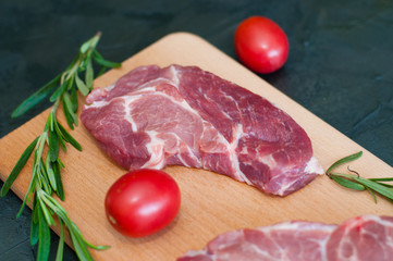 Raw pieces of pork, rosemary and tomatoes on a cutting board, on dark gray concrete background, close-up