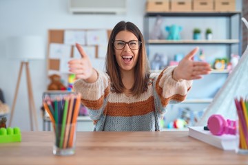 Young beautiful teacher woman wearing sweater and glasses sitting on desk at kindergarten looking at the camera smiling with open arms for hug. Cheerful expression embracing happiness.