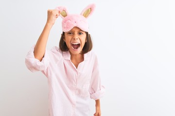 Beautiful child girl wearing sleep mask and pajama standing over isolated white background angry...