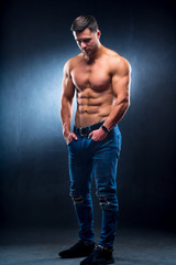 Fototapeta na wymiar Sexual muscular man posing over dark background with naked torso, hands in pockets. Muscular body and strong abs. Looking down. Studio shot.