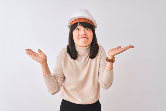 Young beautiful Chinese architect woman wearing helmet over isolated white background clueless and confused expression with arms and hands raised. Doubt concept.