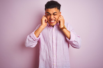 Young brazilian man wearing shirt standing over isolated pink background covering ears with fingers with annoyed expression for the noise of loud music. Deaf concept.