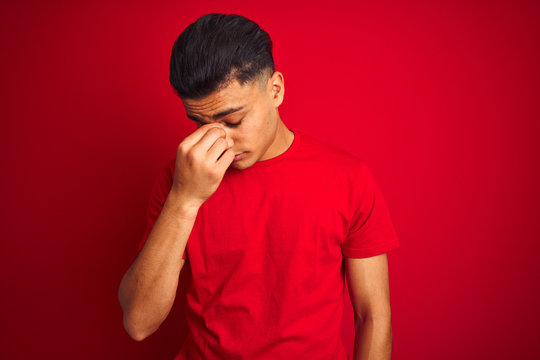 Young brazilian man wearing t-shirt standing over isolated red background tired rubbing nose and eyes feeling fatigue and headache. Stress and frustration concept.