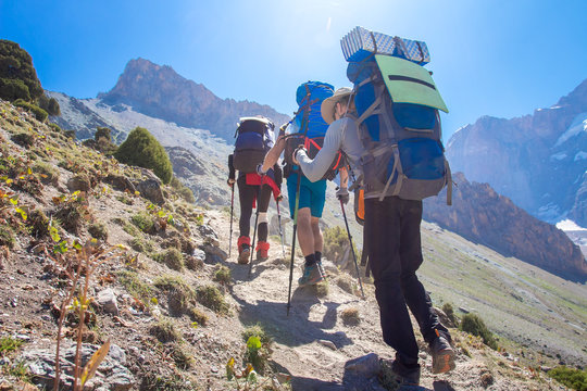 Tourists hiking in mountains with backpacks on sunny summer day. Hikers on mountain trail. Trekking in Fann mountains,  Tajikistan. Climb on mountain summit