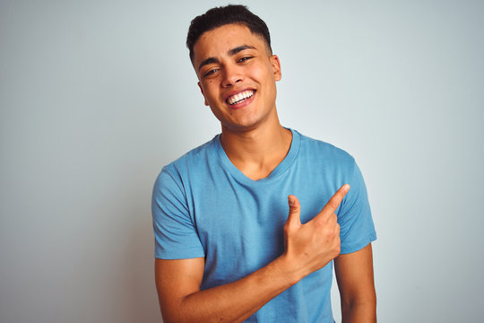 Young brazilian man wearing blue t-shirt standing over isolated white background cheerful with a smile of face pointing with hand and finger up to the side with happy and natural expression on face