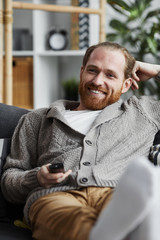 Portrait of modern bearded man smiling happily watching TV at home and holding remote control while...