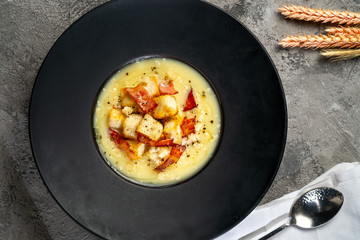 Cream soup with potatoes, bacon and croutons in a black plate on a gray stone background