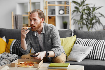 Portrait of middle-aged bearded man watching TV at home and eating pizza during lazy weekend, copy...