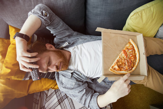 Above View Portrait Of Bearded Man Lying On Couch And Eating Pizza While Watching TV At Home, Copy Space