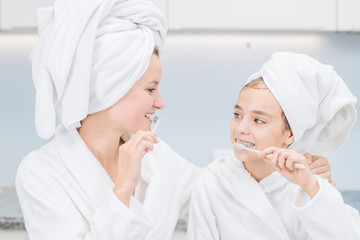 Mother and her daughter are brushing teeth with toothbrushes in the bathroom at home. Mom and child girl are in bathrobes and with towels on their heads
