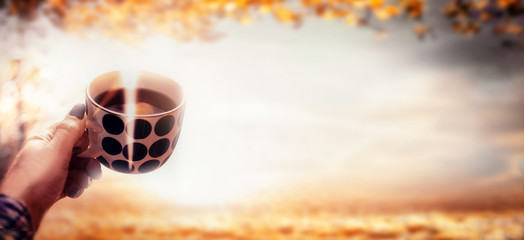 Fototapeta na wymiar Woman hand holding polka dot cup with steamed hot coffee at autumn nature landscape with fall foliage and sunlight. Outdoor. Banner