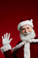 Waist up portrait of classic Santa Claus waving at camera while standing against red background in studio, copy space