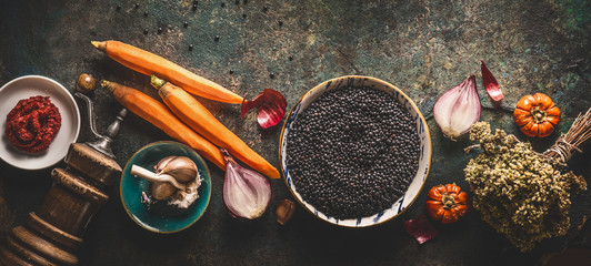 Black lentils with cooking ingredients, vegetables, herbs and spices for tasty vegan meals on dark background. Top view. Healthy vegetarian eating concept. Horizontal banner. Plant based protein - obrazy, fototapety, plakaty