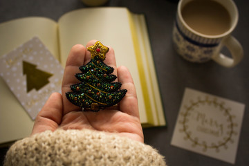 Exclusive Christmas tree beading brooch handmade as an idea for a Xmas gift for a special woman. Collage with woman's hand with jewelry, coffe and note on grey background