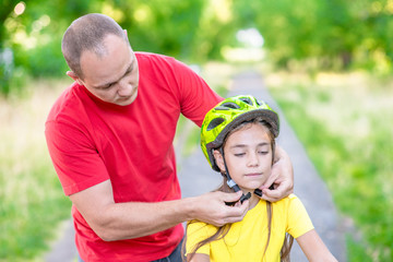 Naklejka premium Father puts a helmet on his daughter in summer park