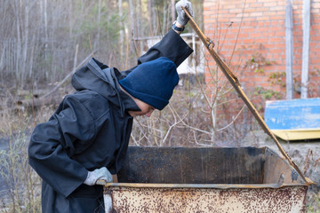 homeless woman in black coat and blue hat is open street garbage cans. woman is rummaging for some food in garbage. poverty.