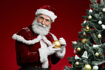 Portrait of cheerful Santa holding golden ornament standing by Christmas tree on red background, copy space