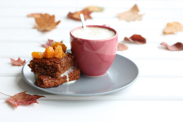 a slice of chocolate cake apricot filling on a plate next to a cup of foamy cappuccino on a table with fallen leaves. sweet and warming autumn coffee pause