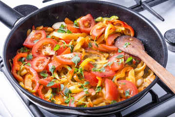 Sliced bell peppers with tomatoes and basil leaves are fried in a pan on a gas stove and mixed with a wooden spoon - cooking homemade sauce