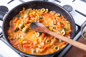 Sliced bell peppers with tomatoes, green olives and basil leaves are cooked in a pan on a gas stove and mixed with a wooden spoon - making homemade Italian sauce
