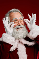 Obraz premium Head and shoulders portrait of classic Santa Claus smiling and adjusting glasses while looking up against red background