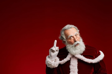 Waist up portrait of surprised Santa Claus pointing up while posing against red background in studio, copy space