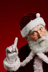 Portrait of classic Santa Claus pointing up while standing against red background in studio