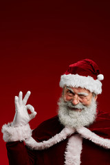 Waist up portrait of winking Santa Claus looking at camera and showing OK sign while posing against...