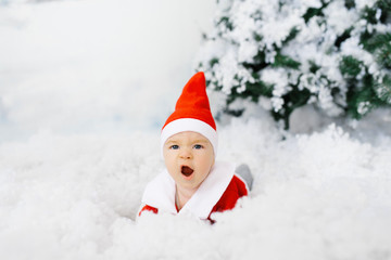 The kid in the clothes of little Santa is lying on his stomach on the artificial snow, he is surprised. Christmas sales concept for kids
