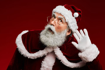 Waist up portrait of classic Santa Claus putting hand to ear trying to hear secrets while standing against red background in studio