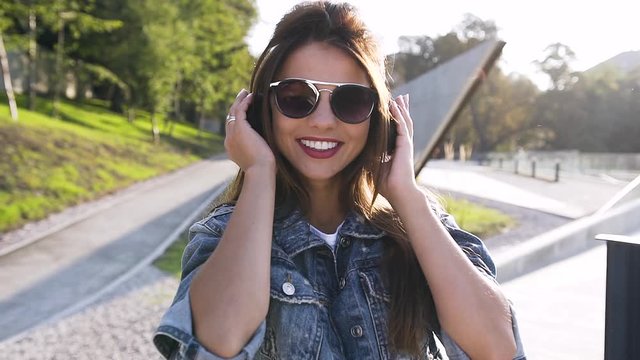 Portrait of pretty young smiling and happy woman in sunglasses with long hair listens music in headphones outdoors on sunny summer day