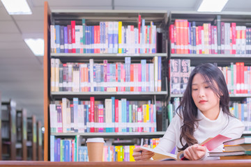 Young beautiful Asian female student portrait sitting and concentrate studying or reading textbook in university library for exams or test with book shelves background. Education and learning concept