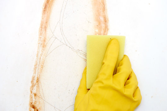 Hand in a yellow glove with a sponge washes rust with a white enamelled bath close up.