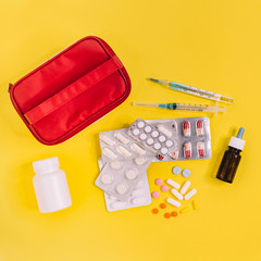 Red home medical cabinet. Blisters with different multicolored pills, syringe, thermometer, nasal drops, white bottle with pillules. First aid kit. Health care concept. Yellow background. Square photo