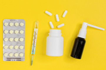 Pills for flu or cold, thermometer, white pillules from bottle and spray for the throat. Minimum set of medicines in the home first aid kit. Healthcare concept. Yellow background, flat lay, close up.