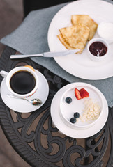 On the wrought iron table stay white plate with pancakes and jam, porridge with fresh strawberry and blueberry and mug of black coffee with cookie. Breakfast in hotel. Selective focus. Vertical photo.