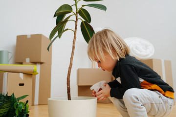 Child watering plant in a room with sealed boxes. His family recently moved in.