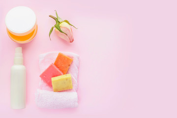 Open jar of cosmetic cream, white cosmetics bottle, soap, rose and towel on pink background. Concept of natural spa cosmetics. Flat lay, top view, copy space .