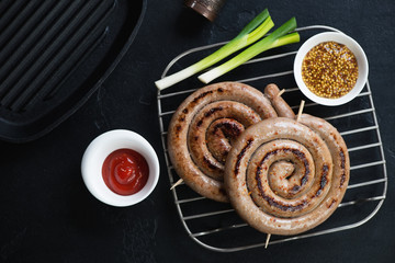 Grilled rolled sausages with ketchup and mustard on a metal cooling rack, top view over black stone...