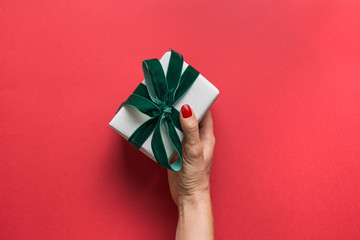 Woman holding Christmas giftbox with green velvet bow on red background. Greeting Xmas card. Boxing day. View from above.