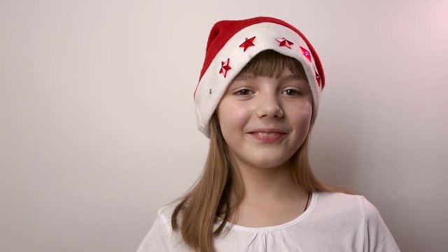eight-year-old funny caucasian girl in Santa Claus hat with blinking red lights in form of stars smiles, laughs on white background. new year or christmas celebration in costume