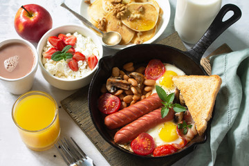 Keto breakfast: fried eggs, sausages, beans, bacon, mushrooms, grilled toasts, orange juice, cottage cheese, pancakes and cocoa on a light stone table.