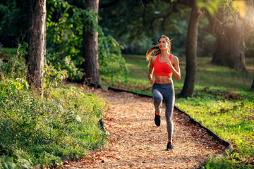 Naklejka na ściany i meble Full length of attractive fit Caucasian brunette in sportswear and with ponytail running on trail in woods. Sunny morning in nature.