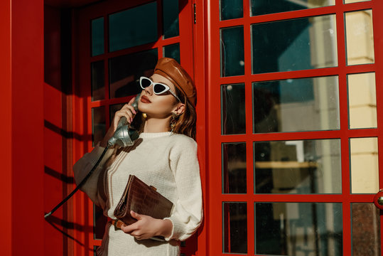 Outdoor Autumn Fashion Portrait Of Young Elegant Woman Wearing Stylish  Beret, Sunglasses, White Sweater, Holding Brown Textured Crocodile Leather Handbag, Posing In Red Call Box. Copy, Empty Space